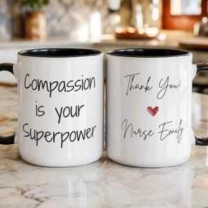 Pode incluir: Duas canecas de cerâmica brancas com bordas e alças pretas. Uma caneca diz "Compassion is your Superpower" em escrita preta. A outra caneca diz "Thank You! Nurse Emily" com um coração vermelho.
