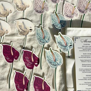 Puede incluir: Tarjetas de lugar con forma de flor, con nombres como "Madison" y "Clarice". Las tarjetas presentan ilustraciones de estilo acuarela de flores de anturio en blanco, morado y verde azulado, con tallos verdes. También se ve un menú.