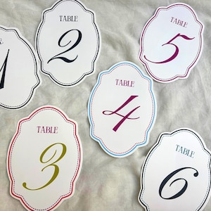 May include: White table number cards with a scalloped edge and the word "TABLE" at the top. Numbers 1 through 6 are displayed in various colors, including black, purple, and gold. The cards are on a white surface.