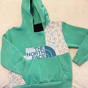 Puede incluir: Una sudadera con capucha verde con paneles estampados florales y un bolsillo delantero. El texto "THE NORTH FACE" está impreso en la parte delantera. La sudadera tiene una capucha con cordón y puños acanalados.