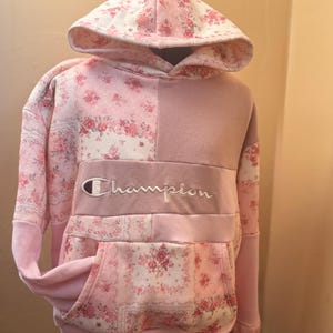 May include: A pink Champion hoodie with a floral pattern on the hood, sleeves, and front pocket. The hoodie features the Champion logo embroidered in white across the chest. The design combines solid pink panels with the floral print.