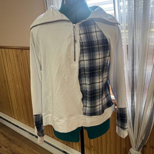 May include: A white long-sleeved sweatshirt with a half-zip collar. The right side and cuffs feature a blue, black, and white plaid pattern. The sweatshirt is displayed on a green dress form.