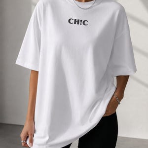 Puede incluir: Una camiseta blanca de manga corta con cuello redondo que presenta el texto negro CH!C impreso en el centro del pecho. La prenda tiene un corte oversize con hombros caídos y un dobladillo recto que cae por debajo de la cintura.