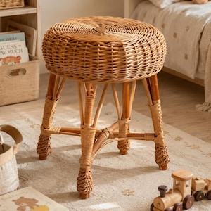 May include: A round stool constructed from light brown woven rattan with a circular seat and four legs reinforced by a central cross-brace. The legs feature decorative braided rattan detailing at the base and mid-sections, providing a stable structure.