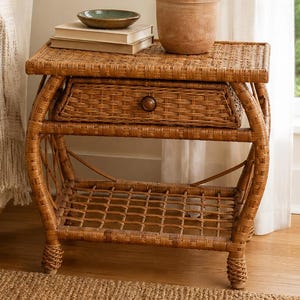 May include: A rectangular brown woven wicker side table featuring a single drawer with a small round wooden knob and an open lower shelf. The table surface holds two stacked books, a small teal bowl, and a potted green plant in a clay pot.
