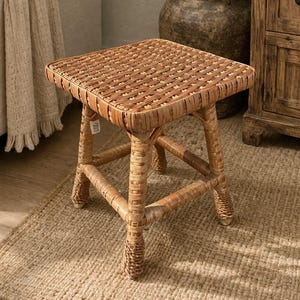 May include: A square, woven rattan stool with a natural finish. The stool has a woven top and four sturdy legs. The stool is placed on a jute rug. The stool is a decorative accent piece for a living room or bedroom.