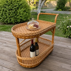 May include: A two-tiered oval serving cart made of woven light brown wicker. The top tier holds a glass dome cover over a wooden platter with cheese, crackers, and grapes. The bottom tier contains two glass wine bottles with labels.