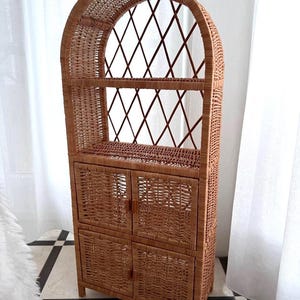 Puede incluir: Un mueble alto de mimbre marrón con una parte superior arqueada y detalles de enrejado de diamantes. El mueble tiene dos estantes y cuatro compartimentos de almacenamiento con puertas. El diseño tejido y el color natural le dan una estética rústica y vintage.