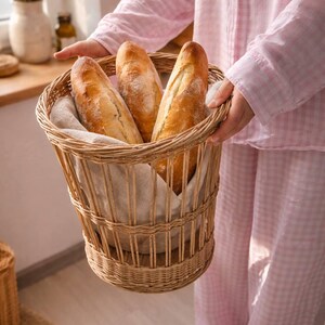 May include: A woven wicker basket holds three fresh baguettes. The basket is lined with a linen cloth. The baguettes have a golden-brown crust. The basket is being held by a person wearing pink pajamas.