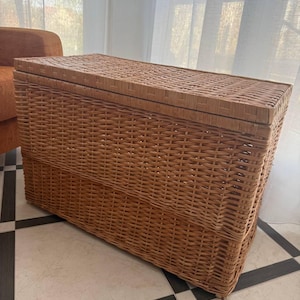 May include: A rectangular, woven wicker storage trunk with a hinged lid. The trunk is a warm, natural brown color and features a tightly woven pattern. It is placed on a tiled floor with a black and white checkered design. The background includes a window with sheer white curtains and an orange armchair.