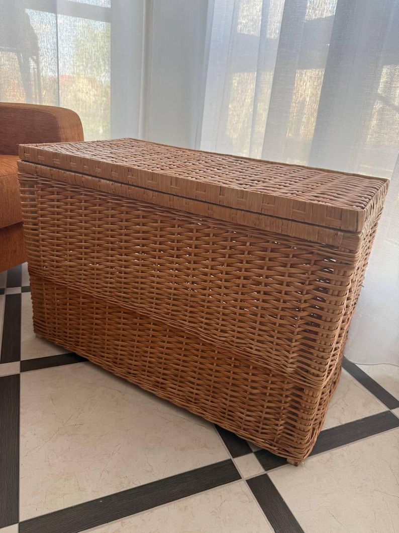 May include: A rectangular, woven wicker storage trunk with a hinged lid. The trunk is a warm, natural brown color and features a tightly woven pattern. It is placed on a tiled floor with a black and white checkered design. The background includes a window with sheer white curtains and an orange armchair.