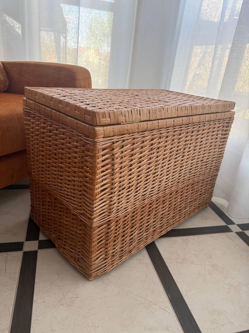 May include: A rectangular wicker storage trunk with a hinged lid. The trunk is woven from light brown wicker and has a textured surface. The trunk is placed on a tiled floor and is next to a window with white curtains.