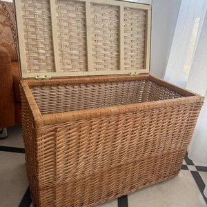 May include: A rectangular wicker storage chest with a hinged lid, open to reveal the interior. The chest is made of woven light brown wicker, with a lighter-colored wooden frame on the lid. The chest is sitting on a tiled floor.
