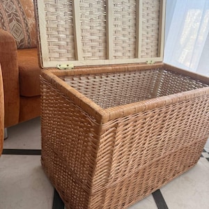 May include: A rectangular wicker storage chest with a hinged lid. The chest is made of woven light brown wicker and has a rectangular shape. The lid is open, revealing the interior. The chest is placed on a tiled floor.