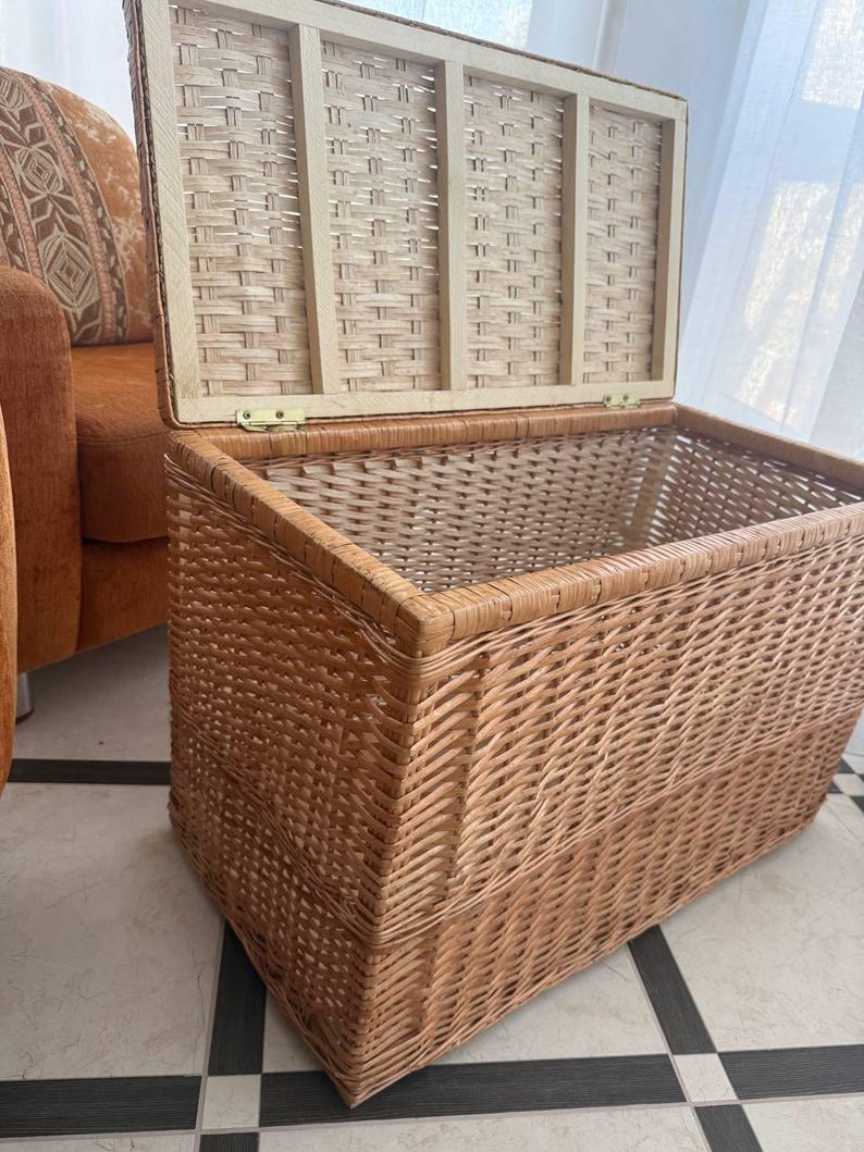May include: A rectangular wicker storage chest with a hinged lid. The chest is made of woven light brown wicker and has a rectangular shape. The lid is open, revealing the interior. The chest is placed on a tiled floor.