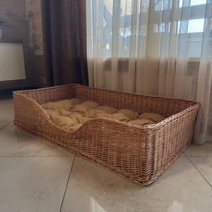 Könnte beinhalten: Ein rechteckiges Haustierbett aus geflochtenem Korb in Naturbraun. Das Bett hat ein weiches, hellbraunes Kissen im Inneren. Das Bett steht auf einem gefliesten Boden, mit einem Fenster und Vorhängen im Hintergrund.