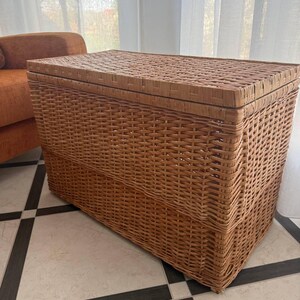 May include: A rectangular woven wicker storage trunk with a hinged lid. The trunk is a light brown color and has a textured surface. The trunk is sitting on a tiled floor.