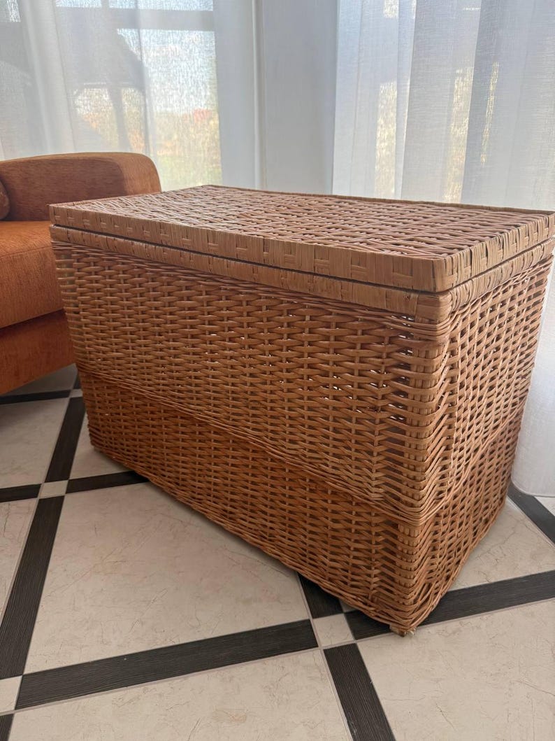 May include: A rectangular woven wicker storage trunk with a hinged lid. The trunk is a light brown color and has a textured surface. The trunk is sitting on a tiled floor.