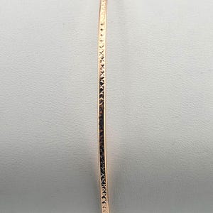 Puede incluir: Una pulsera brazalete de oro rosa con un diseño texturizado y martillado. La pulsera fina y redonda se muestra sobre un fondo blanco. La superficie de la pulsera tiene un patrón repetitivo de pequeñas hendiduras anguladas.