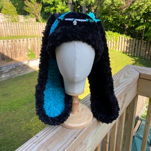 May include: A black plush bunny hat with long, floppy ears. The interior of the ears is bright turquoise. The hat is decorated with a beaded headband and a small skull charm. A turquoise bow is attached to the side.
