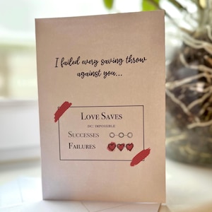 May include: A folded greeting card with the text "I failed every saving throw against you..." and "Love Saves." The card features a game-like design with hearts and circles, and red brush strokes. The card is tan.