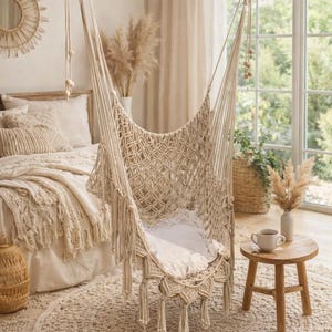 May include: A beige macrame hammock chair hangs from a wooden beam, with a white cushion inside. The chair is suspended above a beige, circular rug. The room has a neutral color palette, with a bed and a small wooden side table.
