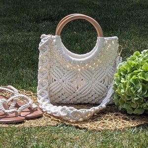 May include: A cream-colored macrame handbag with wooden handles and a matching strap sits on a woven mat. Beside it are cream-colored sandals with brown soles. A green plant is to the right, all on a grassy background.