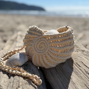 May include: A cream-colored macrame bag featuring a spiral knot pattern on the side, accompanied by a gold-toned chain strap. The bag is adorned with two white seashells, one placed inside the opening and one resting near the chain strap.