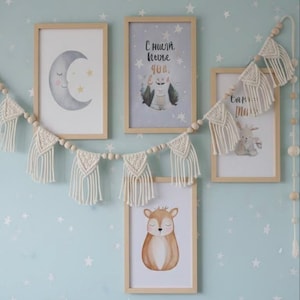 May include: A collection of framed art prints and a macrame garland. The prints feature watercolor illustrations of a crescent moon, a deer, and a creature with text. The garland is made of cream-colored macrame and wooden beads, creating a cozy, whimsical aesthetic.