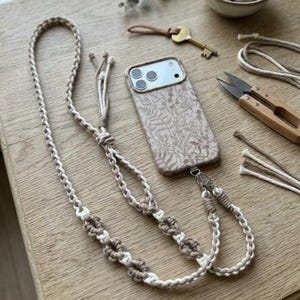 May include: A beige phone case with a floral pattern and a matching macrame phone strap. The strap is made of braided beige and white cord with decorative knots. The phone case has a silver-colored clasp.