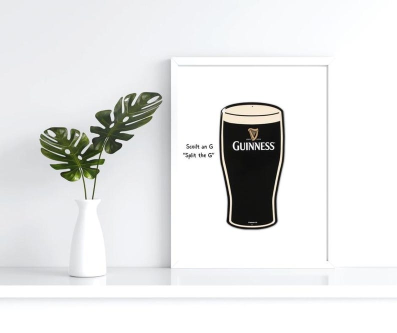 May include: A framed print featuring a pint of Guinness stout. The black and white image has the Guinness logo and text that reads "Scoilt an G Split the G". A white vase with green leaves is on the left.