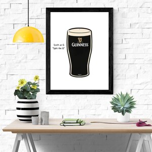 May include: A framed print featuring a stylized pint of Guinness stout. The black and white image has the Guinness logo and the text "Scoilt an G Split the G". The print is in a black frame and is displayed on a white brick wall.