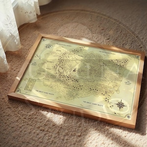 May include: A framed map of Oz, featuring detailed illustrations of the land and its regions. The map is set against a light yellow background, with a compass rose in the lower right corner. The frame is made of light-colored wood.