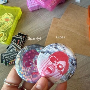 May include: Two circular pin-back buttons featuring a disco ball pattern background. The left button has a sparkly finish, while the right button has a gloss finish. Both buttons display a red-toned graphic of a person with hands on their face.