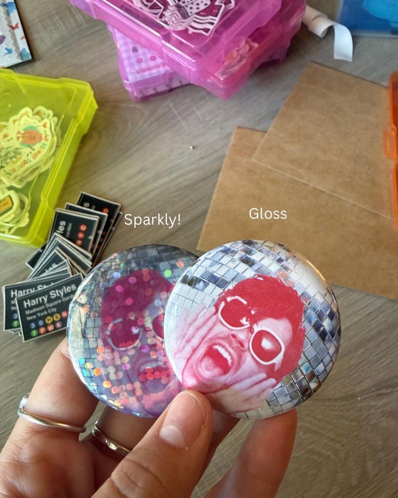 May include: Two circular pin-back buttons featuring a disco ball pattern background. The left button has a sparkly finish, while the right button has a gloss finish. Both buttons display a red-toned graphic of a person with hands on their face.