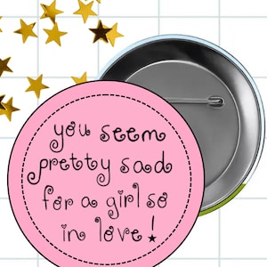 May include: A round, silver-backed button with a pink front. The pink front has a black dotted border and black text that reads "you seem pretty sad for a girl so in love!" with a small star at the end.