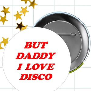 May include: A round pin-back button featuring a white background with the red text "BUT DADDY I LOVE DISCO" printed in a bold, serif font. The back of the button is metallic silver with a horizontal pin mechanism for attachment to clothing.