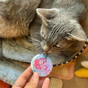 May include: A circular pinback button featuring a pink skull graphic set against a blue and purple iridescent background with embedded holographic glitter. The design has a glossy finish and a grid-like pattern overlaying the skull illustration.