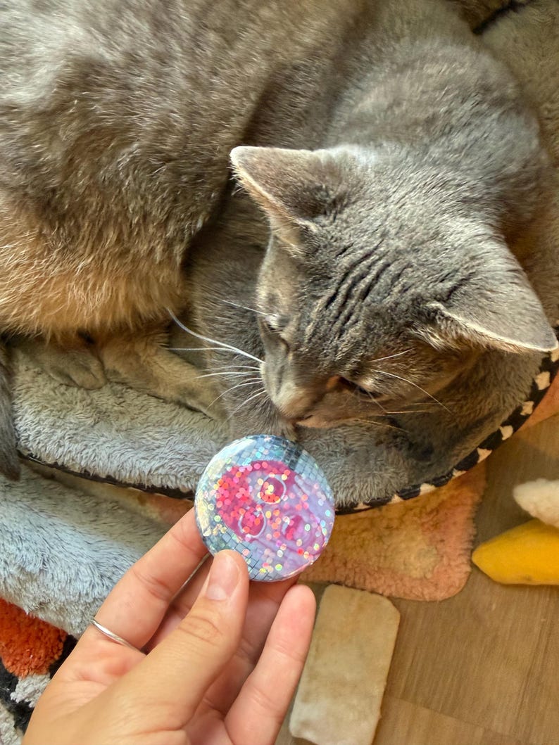 May include: A circular pinback button featuring a pink skull graphic set against a blue and purple iridescent background with embedded holographic glitter. The design has a glossy finish and a grid-like pattern overlaying the skull illustration.