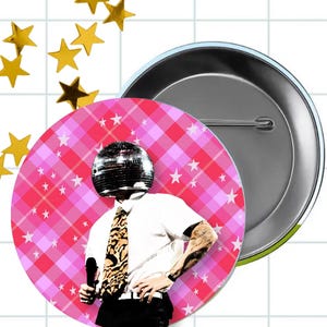 May include: A circular pin-back button featuring a graphic of a person with a disco ball head, wearing a white short-sleeved shirt and a leopard print tie. The background is a pink and white plaid pattern decorated with small white stars.