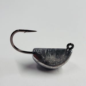 May include: A silver-colored fishing jig head with a curved, textured body and a black hook. The hook extends from the body, with a loop at the top for attaching to fishing line. The jig head is designed for fishing.
