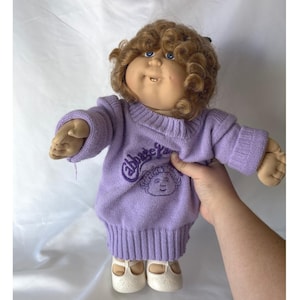 May include: A vintage Cabbage Patch Kid doll with light brown curly hair and blue eyes. The doll wears a lavender sweater with the name "Abbey" and a matching image. The doll has a soft, fabric body and is wearing white shoes.