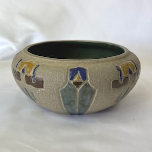 May include: A round, beige ceramic bowl with a dark green interior. The exterior features a repeating pattern of stylized geometric shapes in blue, yellow, and brown. The bowl has a textured surface.