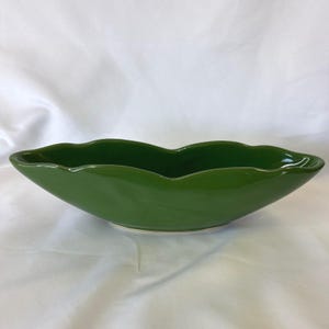 May include: A glossy, green ceramic serving bowl with a scalloped rim. The oval-shaped bowl has a smooth, reflective surface and a white base. The bowl is empty and sits on a white background.