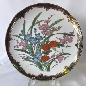 May include: White ceramic decorative plate with a scalloped edge, featuring a floral design with blue, pink, and orange flowers. The rim has a gold border and a dark blue patterned edge.