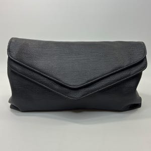 May include: A black envelope-style clutch bag featuring a textured leather-like surface. The bag has a pointed flap closure that extends across the front, creating a distinct V-shaped edge. The overall design is rectangular with a soft, flexible structure.