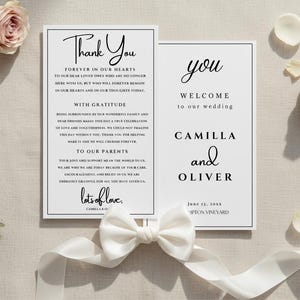 May include: Elegant wedding invitation cards with floral accents. The cards feature the words "Thank You" and "Welcome" with the names Camilla and Oliver. A white bow and ribbon are placed near the cards.