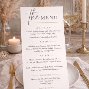 Könnte beinhalten: Eine Hochzeitsmenükarte mit dem Text "The Menu" in eleganter Schrift. Die Speisekarte listet Vorspeisen, Salat, Hauptgerichte und Dessertoptionen auf. Die Karte befindet sich auf einem Tisch mit einer goldenen Gabel und einem Messer, einer brennenden Kerze und einem Glas Wein.