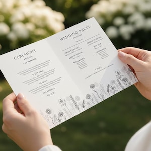 Puede incluir: Un programa de boda con texto negro elegante e ilustraciones florales. El programa se sostiene abierto, mostrando los detalles de la ceremonia y la información de la fiesta de bodas. El fondo es un verde suave y borroso.