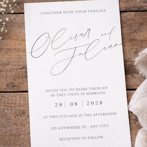 Puede incluir: Una invitación de boda blanca con una escritura elegante. La invitación dice "Oliver y Julian" e incluye la fecha "28 | 08 | 2028". El texto es negro, con un toque de oro. La invitación está sobre una superficie de madera.